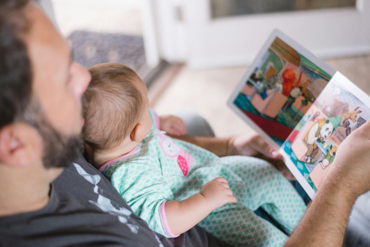 Papà con in braccio suo figlio neonato mentre gli legge un libro sul divano